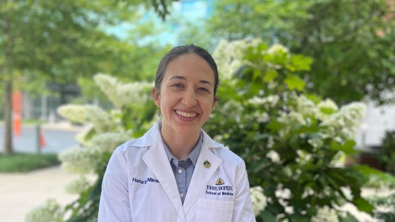 Hana Minsky in a white Johns Hopkins lab coat smiling with greenery in the background