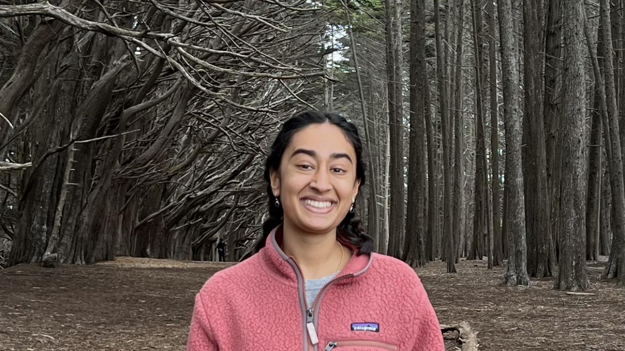 Maya wearing a pink jacket and smiling at the camera in a forest