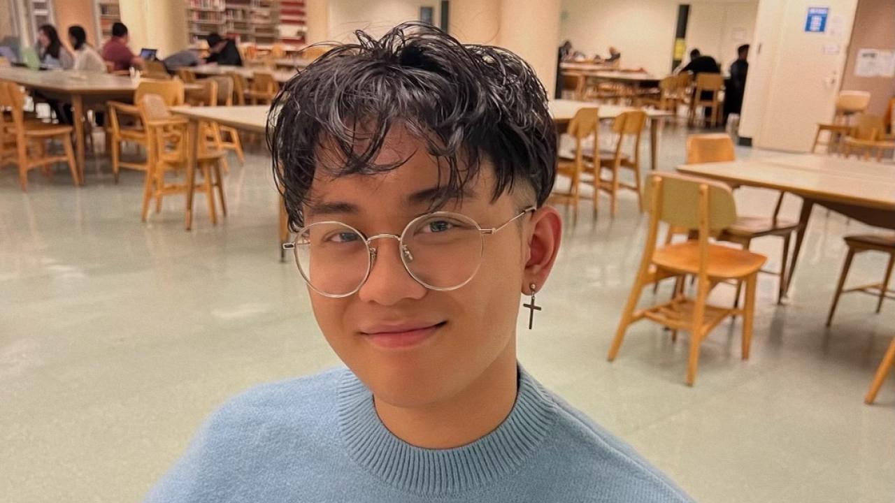 robert chin smiling at the camera wearing a duck sweater and sitting in the library