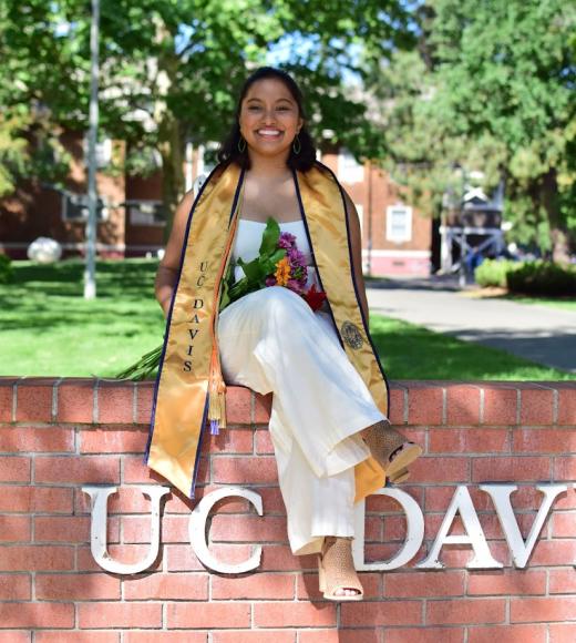indira wearing a UC Davis stole and smiling on top of the brick UC Davis sign holding flowers
